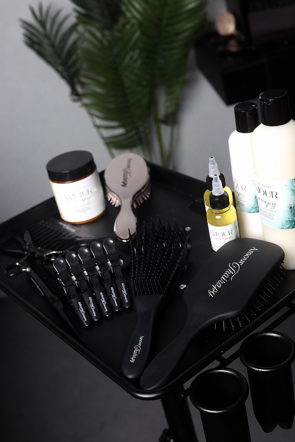A Amour Thairapy haircare and styling set arranged on a black tray, including a large black hairbrush, several smaller black brushes, a small yellow bottle, two white bottles with black caps, and a white jar with a black lid. The products are photographed in a styled indoor setting with a dark background and blurred green leaves.