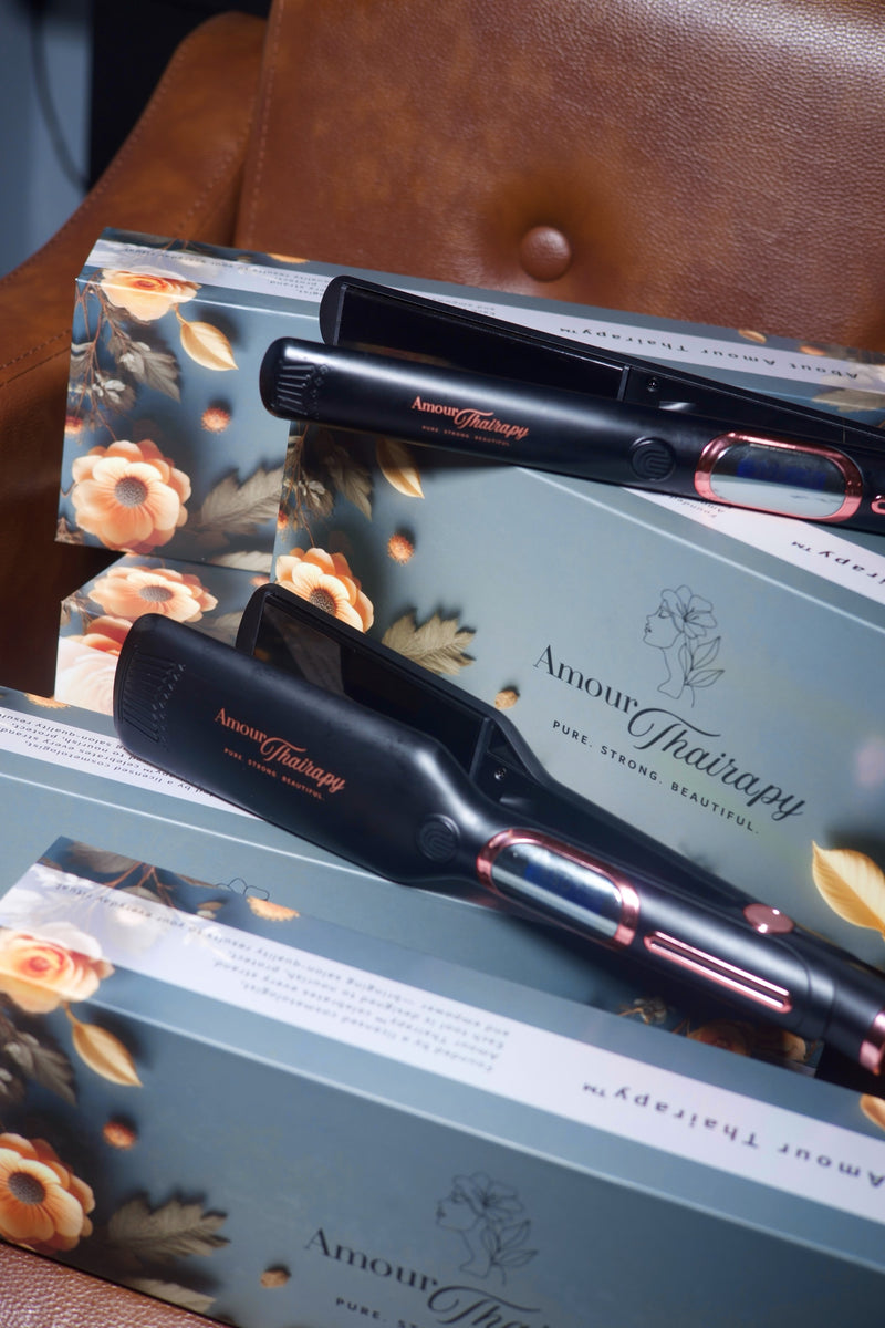 Two black hair straighteners with rose gold accents resting on top of their floral-patterned Amour Hairistry packaging, placed on a brown leather surface.