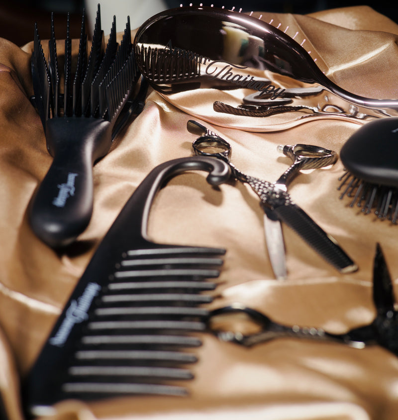 A close-up flat lay of black haircare tools and accessories arranged on shiny champagne satin, including a wide-tooth comb, paddle brush, hair clips, and scissors. The brand name 'Chicory' is visible on several items.