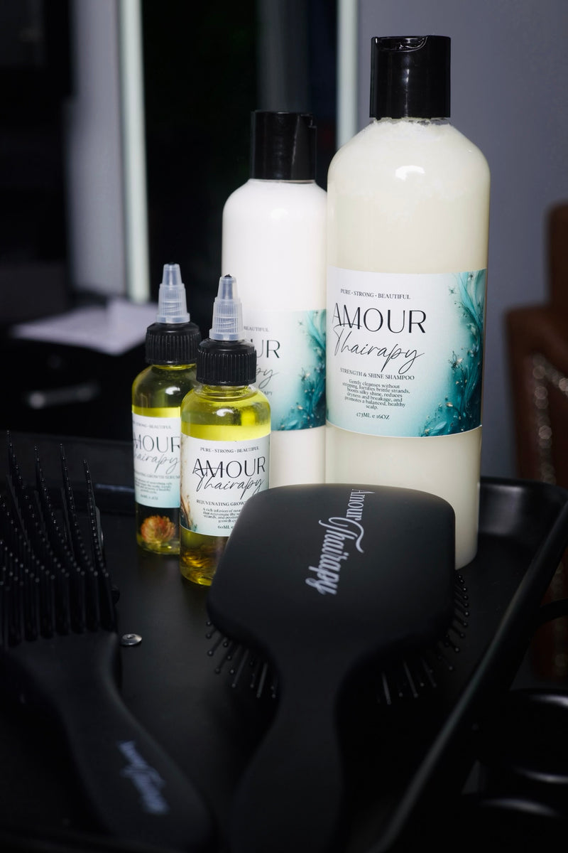 A set of Amour Hairculty hair care products on a black tray, including a large white bottle of shampoo, two small dropper bottles of treatment, and a black paddle hairbrush.