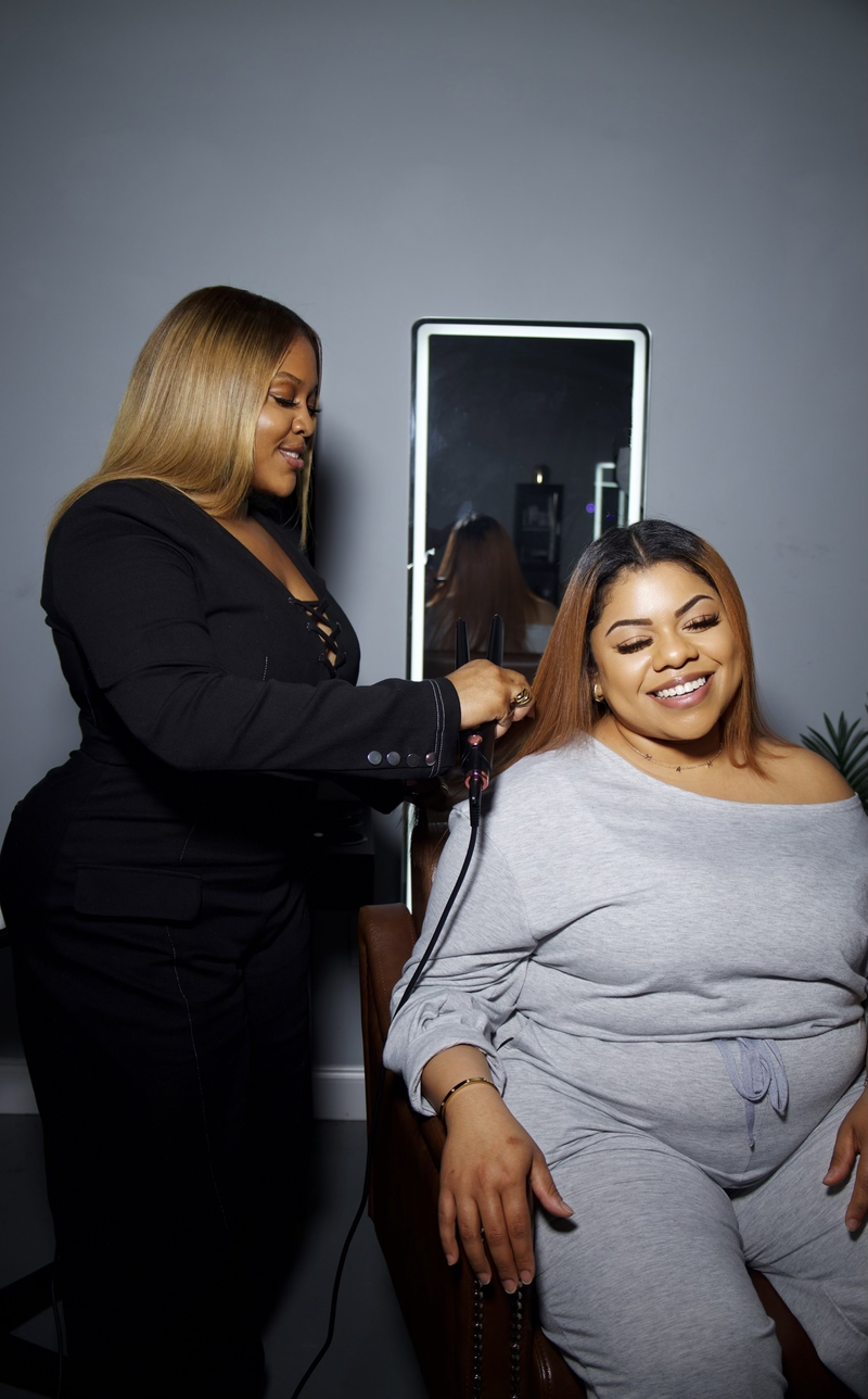 A woman in a black outfit styles the hair of another woman wearing a light gray off-the-shoulder sweatshirt and matching sweatpants, seated in a salon chair with a mirror behind them.